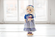 Portrait Of Textile Handmade Vintage Doll With Blue Eyes, Long Blond Hair In Old Blue Textile Dress With Gentle Print, In White Shirt With Purple Boots On White Background. She Holding Little Textile