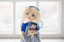 Portrait Of Textile Handmade Vintage Doll With Blue Eyes, Long Blond Hair In Old Blue Textile Dress With Gentle Print, In White Shirt With Purple Boots On White Background. She Holding Little Textile