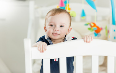 Wall Mural - Portrait of cute baby boy standing in white wooden crib and looking in camera