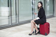 Manager vestita con un tailleur nero con il cellulare in mano è seduta sopra una valigia rossa sta aspettando spazientita- sfondo corridoio esterno in marmo