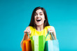 Happy smiling brunette girl wears yellow shirt holding colorful shopping bags on blue background, shopaholic sale concept