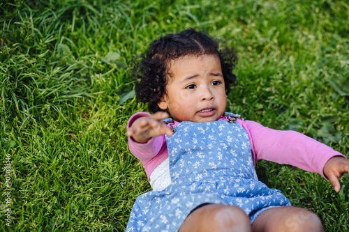 Fotografia, Obraz Dark skinned hispanic toddler girl reaching hand for help with hand while lying on the grass in the summer day in park