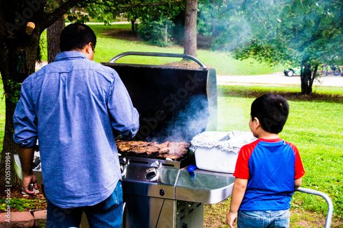 Fotografie, Obraz Latino dad cooking on a grill outside while son watches