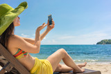 Fototapeta  - Attractive young woman sending online a love message through a selfie photo on the mobile phone while sitting on the beach during summer vacation