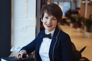 Beautiful Young cute girl sitting in a blue jacket at a table in a cafe, reading a book, looking at the camera, smiling