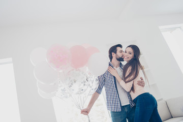 Wall Mural - Low angle view portrait of attractive positive couple in casual outfits man preparing gift, present for  lover embracing kissing in cheek, standing in modern white apartment