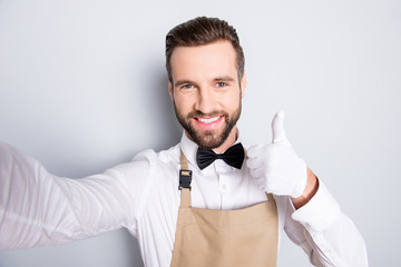 Wall Mural - Self portrait of attractive cheerful man in white shirt black bow, modern hairstyle, shooting selfie on front camera and gesture thumb up isolated grey background, having fun, rest, relax, video-call