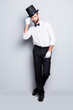 © deagreez - Full size fullbody portrait of stylish attractive magician in tophat, formal wear,  looking at camera, staying leg by foot, holding hat with hand, isolated on grey background