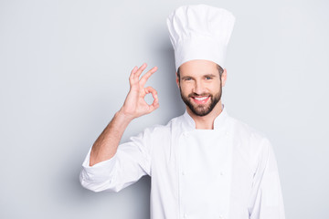 Wall Mural - Portrait of cheerful joyful chef cook in beret and white outfit with stubble looking at camera showing ok sign, approve, advice menu isolated on grey background