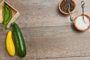 Top view of a wood box of green string beans, freshly picked, raw yellow squash, zucchini and two wood bowls of sea salt and fresh pepper corns with artisan spoons on a wood table