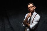 Fototapeta  - Young stylish hipster with cool hairstyle and beard dressed in white shirt and suspenders is thinking of a new creative idea looking at viewer