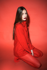 Wall Mural - Woman with stylish makeup and long hair posing in total red outfit. Fashion concept. Girl on calm face in red formal jacket and tights, red background. Lady looking at camera while sitting on floor.