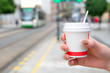 Female hand holding a white paper coffee cup in a tram stop with a tram arriving in the background. Concept of take away coffee and commuting 