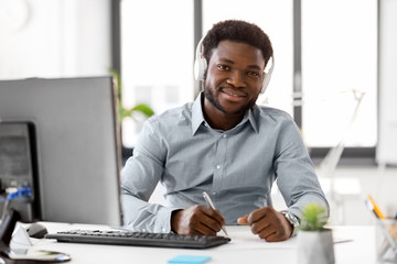 Wall Mural - business, technology and people concept - happy african american businessman with headphones and papers listening to music at office