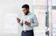 © Syda Productions - business, people and technology concept - african american businessman with smartphone having break and drinking coffee at office glass board