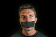 © TheVisualsYouNeed - young man with mouth and lips sealed covered with adhesive tape in censorship coerced freedom of speech and forced silence and secrecy