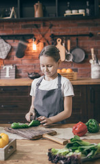 Wall Mural - Young cute girl in apron standing on kitchen and cutting the cucumber. 