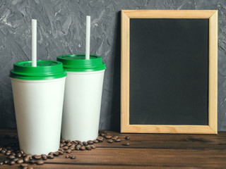 two white plastic coffee cups with a green lid and an inscription frame on a wooden table with scattered coffee beans