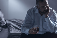 Close-up Of A Man With Midlife Crisis Sitting On A Bed With One Hand On His Chin And Holding A Glass Of Alcohol In The Other