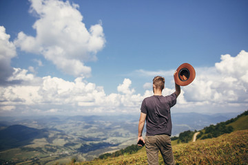 stylish traveler man with hat holding phone, walking on top of sunny mountains and sky. travel and wanderlust concept. space for text. happy hipster traveling. summer vacation