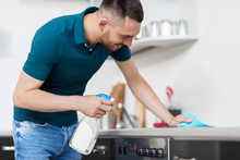 cleaning, household and people concept - man with spray cleaner and cloth wiping cooker at home kitchen