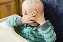 Playful Baby Boy Covering Eyes While Playing Peekaboo On High Chair At Home