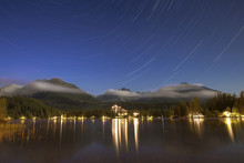 Majestic View Of Lake By Illuminated Town Against Mountains And Star Trails At Night