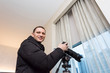 © Kristina Blokhin - Young happy smiling man photographer with tripod taking pictures inside room of hotel, apartment building through window in New York City, NYC urban