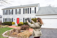 Young Woman, Female Homeowner Standing In Front Of House On Windy Day In Coat Jacket During Storm Holding Up Roof Tile Shingle Inspecting Damage