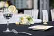 © Kristina Blokhin - Closeup of one glass with lemon slice and water in restaurant or wedding reception black table tablecloth, plate with cake slice, bouquet flowers, fork, chairs, dishes