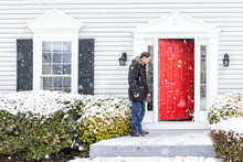 Young Man Outside Front Yard Red Door Of House With Snow During Blizzard White Storm, Snowflakes Falling Letting Calico Cat Outside Outdoors To Porch