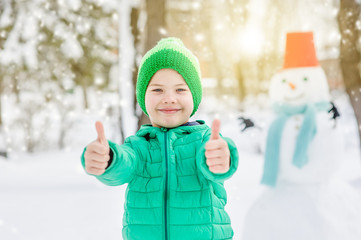 Wall Mural - Smiling boy with snowman on background showing thumbs up