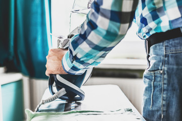 Wall Mural - Man at home ironing clothes. Father performs home duties, irons the child's clothes. Man in role of a woman at home. Man ironing shirt on ironing board with steaming blue iron.