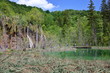 Park Jezior Plitwickich w Chorwacji, szmaragodwa toń wody, wzgórze porośnięte gęstą zielenią, wodospad, turyści podziwiający piękno przyrody, błękitne niebo