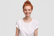 Waist up portrait of delighted freckled redhead woman with pleasant smile, wears casual white t shirt, rejoices good news, isolated over studio background. Foxy female teenager spends time with friend