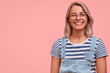 Charming pleasant looking female in stylish denim dungaree and spectacles, listens carefully to somebody`s point of view, expresses positiveness, stands against pink background with copy space