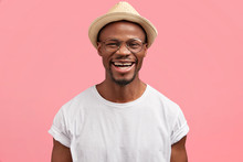 Middle Aged Dark Skinned Hipster In Stylish Hat, Giggles And Smiles Gladfully At Camera, Being In Good Mood After Successfully Prepared Project, Isolated Over Pink Background. People, Happiness