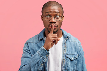 Wall Mural - Secret gesture. Portrait of attractive dark skinned African American male shows silence sign, asks to keep private information confidential, has surprised expression, isolated over pink background.