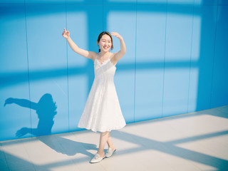 Wall Mural - Beautiful Chinese brunette woman in white wedding dress dancing and smiling at camera against blue wall background. Sunny portrait of glamour young stylish lady. Emotions, beauty and lifestyle concept