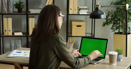 View from behind on the woman sitting at the office table and taping on the keyboard of the laptop computer with green screen, then drinking a coffee. Chroma key. Inside