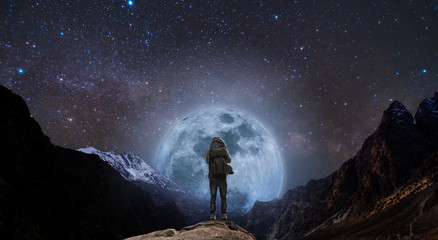 a man with backpack standing on mountain peak at night, and silhouette mountain with bright full moo