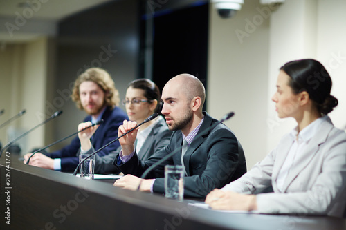 Fotografía One of delegates speaking in microphone while sitting by tribune among other par