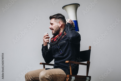 Photographie Side view of young attractive movie director shouting on the megaphone while sitting on the chair isolated on grey background