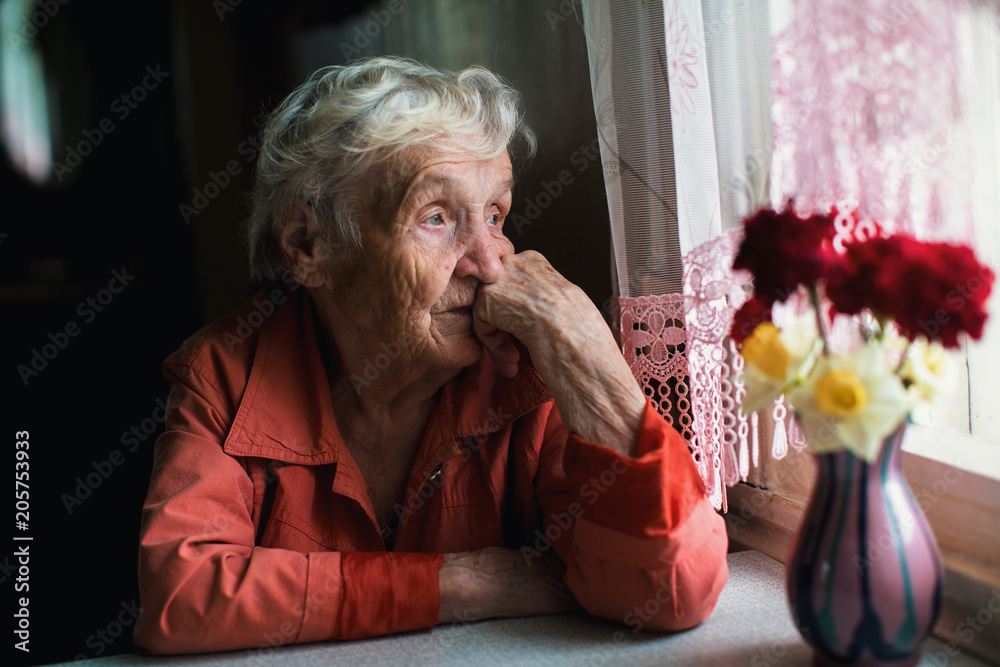 Elderly woman looks sadly out the window. - obrazy, fototapety, plakaty 