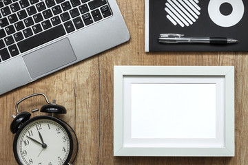 A wooden desk with a laptop, a black watch, a notebook in circles and a white empty frame