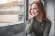 Leinwandbild Motiv Portrait of red-haired happy woman resting at home. She is smiling while looking through window with pleasant expectation. Copy space in left side