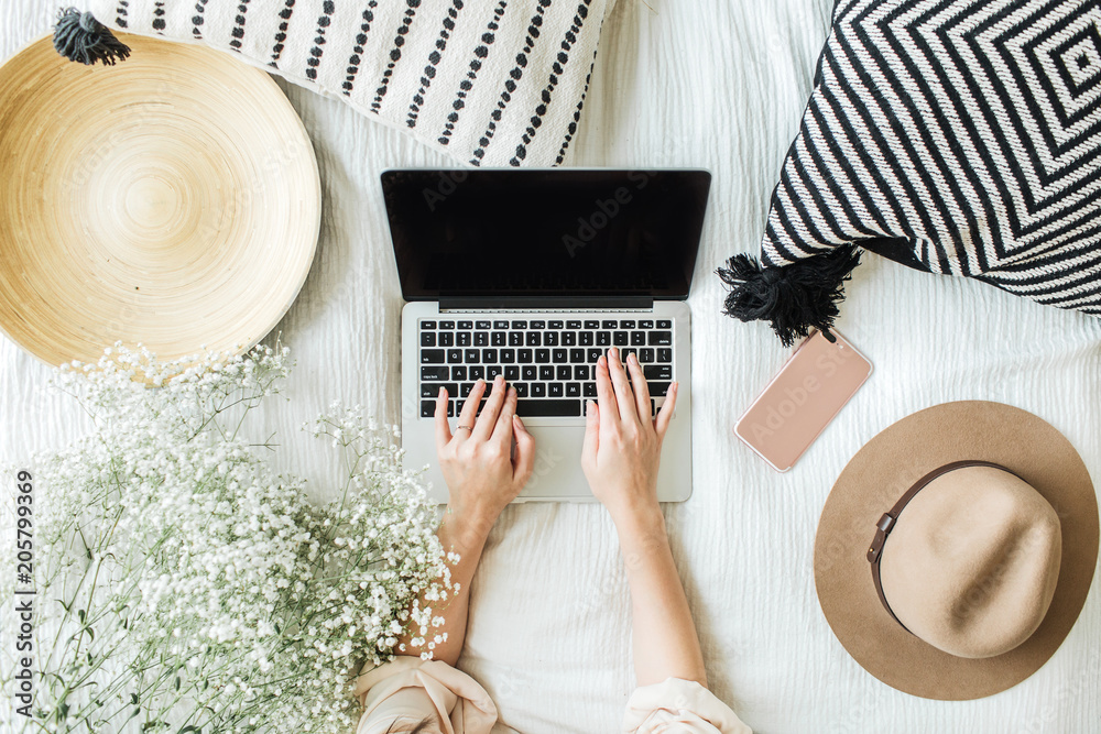 Young woman typing on laptop in bed. Lifestyle hero header with view from above with white flowers bouquet, pillow, hat, cell phone and wooden plate. Freelancer or fashion blogger home workspace - obrazy, fototapety, plakaty 
