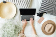 Young woman typing on laptop in bed. Lifestyle hero header with view from above with white flowers bouquet, pillow, hat, cell phone and wooden plate. Freelancer or fashion blogger home workspace