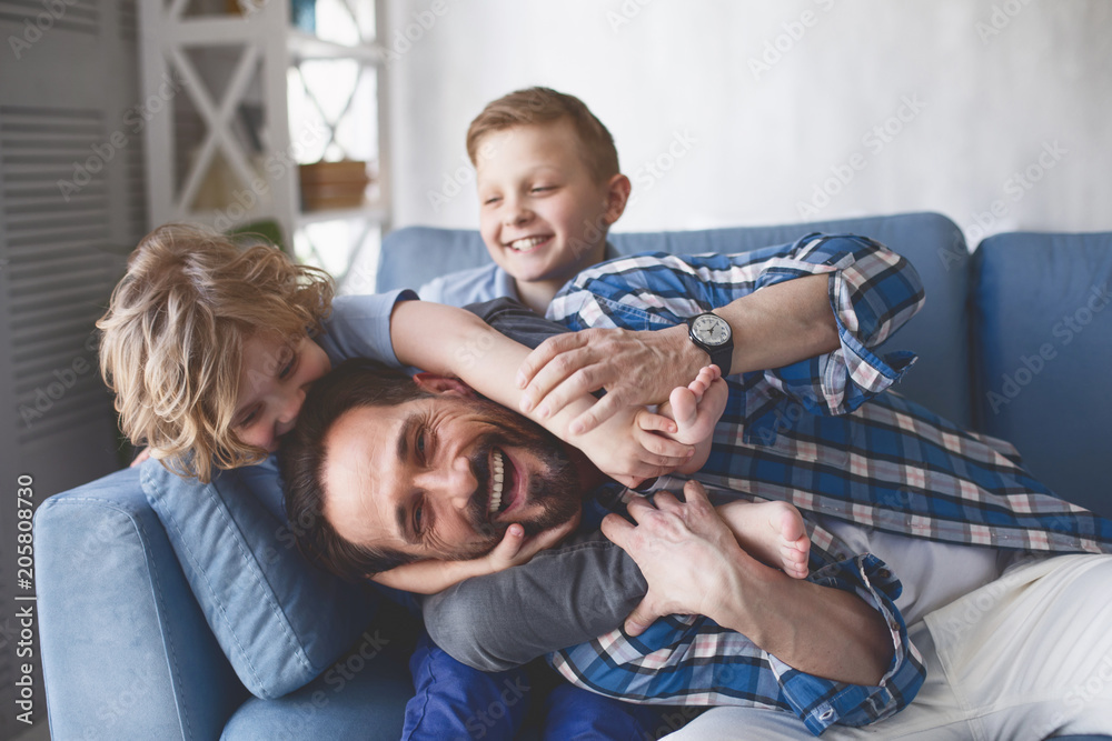 Portrait of smiling father and outgoing sons having fun on cozy couch in apartment. Happy family concept - obrazy, fototapety, plakaty 