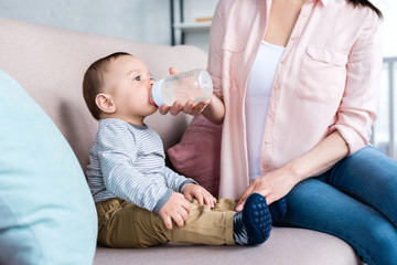 cropped shot of mother feeding her little child with bottle at home
